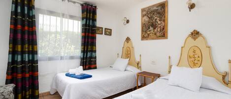A hotel room with two beds, a window with curtains, a painting on the wall, and a small table.