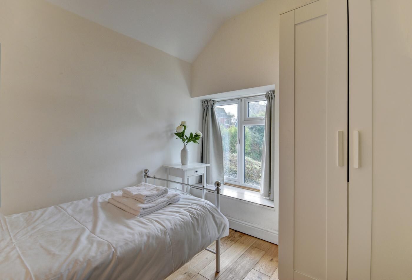 A bedroom with a single bed, a bedside table with a vase of flowers, a window with curtains, and a wardrobe.