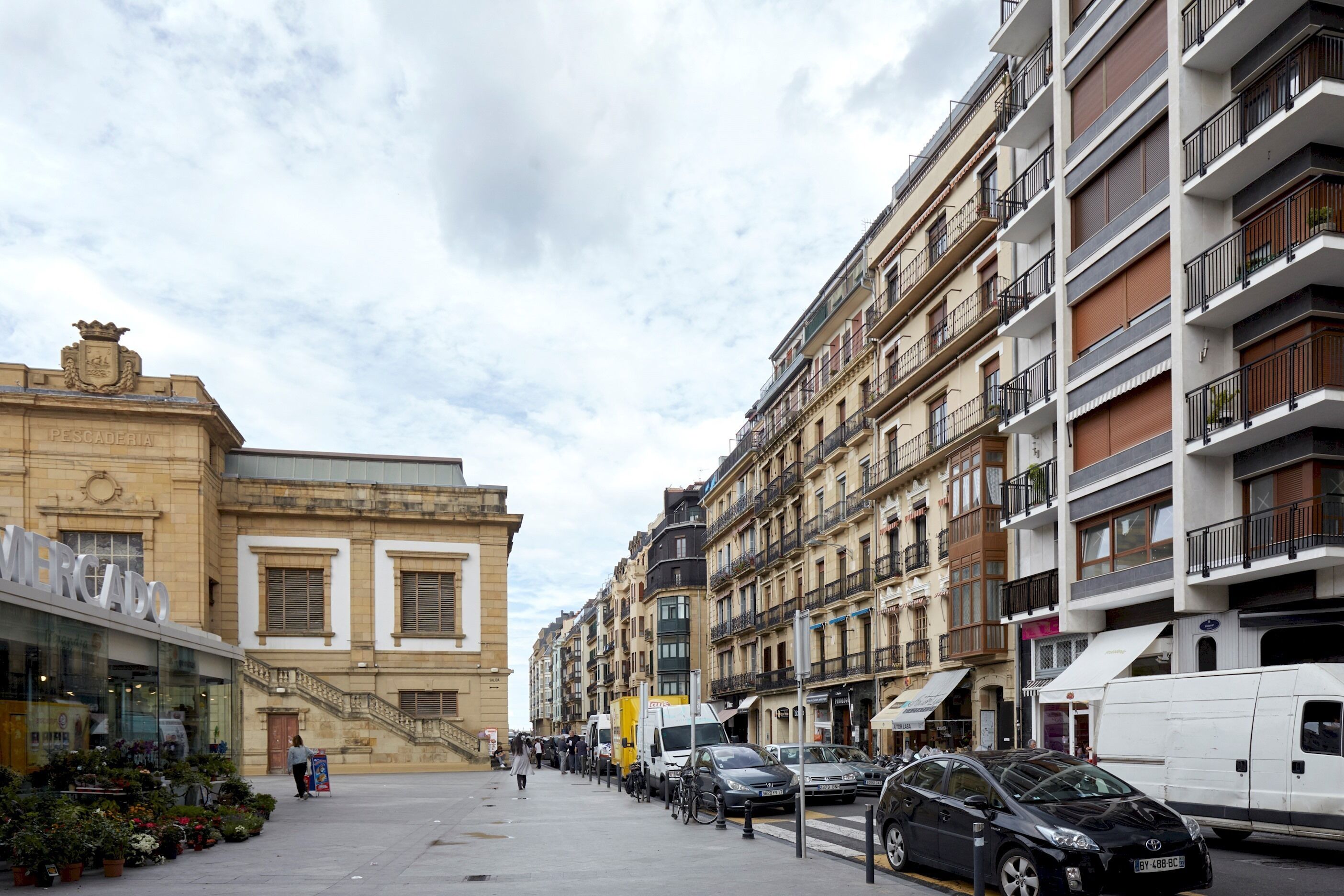 Una calle con edificios, un mercado y coches aparcados.