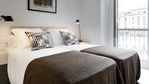 Ein Hotelzimmer mit großem Fenster, einem Bett mit weißer Bettwäsche und gemusterten Kissen, einem Nachttisch mit Lampe und Blick auf die Stadtlandschaft.