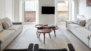Ein modernes Wohnzimmer mit einer Couch, zwei runden Tischen, einem Fernseher und großen Fenstern mit Blick auf Gebäude.