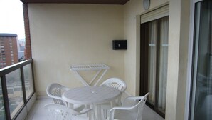 En balkon med et hvidt bord og fire stole, en lille sort genstand på væggen, og udsigt til bygninger gennem glasdøren.