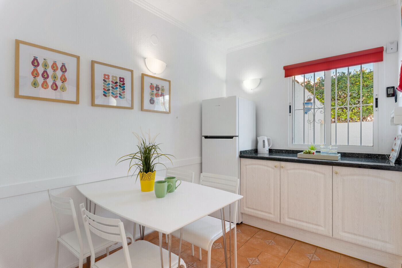 A kitchen with white cabinets, a white refrigerator, a dining table with chairs, and a window with a red blind.