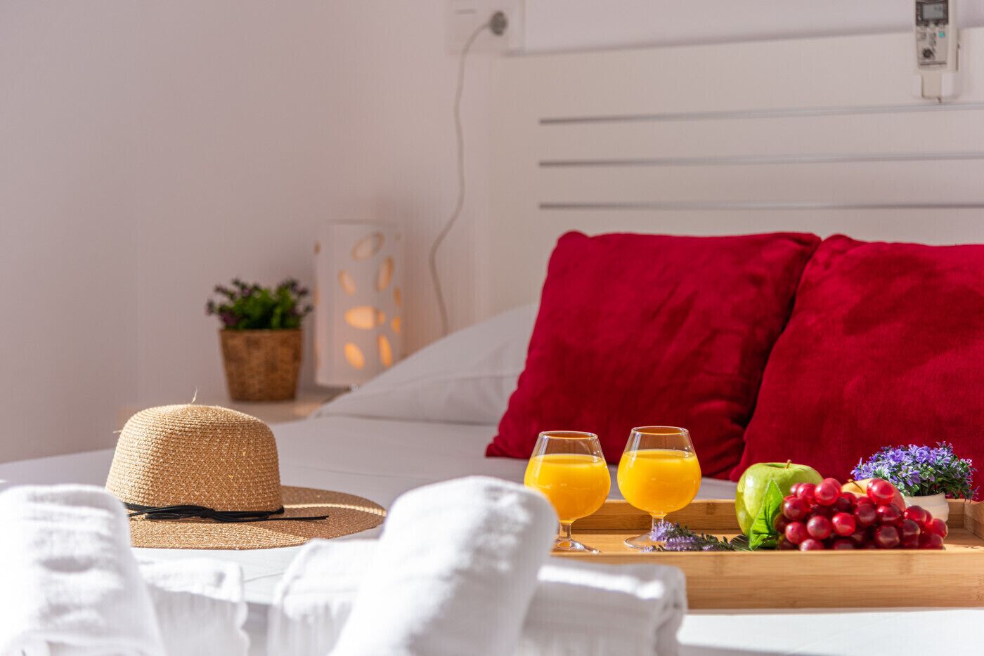 Una cama bien hecha con un almohadón rojo, un sombrero de paja, dos vasos de jugo de naranja y una bandeja con frutas y una pequeña planta.