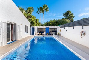 Une piscine avec un espace patio, une cuisine extérieure couverte et un bâtiment blanc avec une porte bleue.