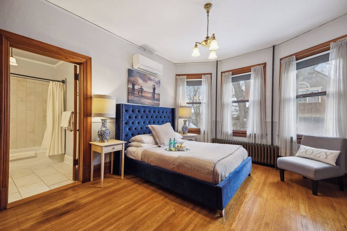 Quarto com cama grande, uma cadeira, mesa de cabeceira, abajur e pintura na parede.