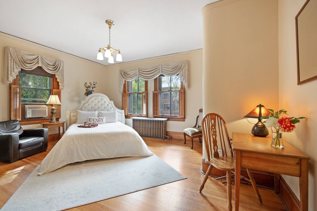 Quarto com uma cama grande, uma escrivaninha de madeira, uma cadeira, um abajur e uma janela com persianas.