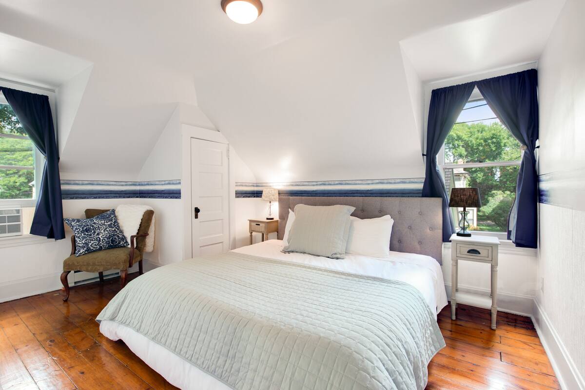 Quarto com uma cama grande, uma cadeira, uma mesa de cabeceira e uma janela com cortinas.
