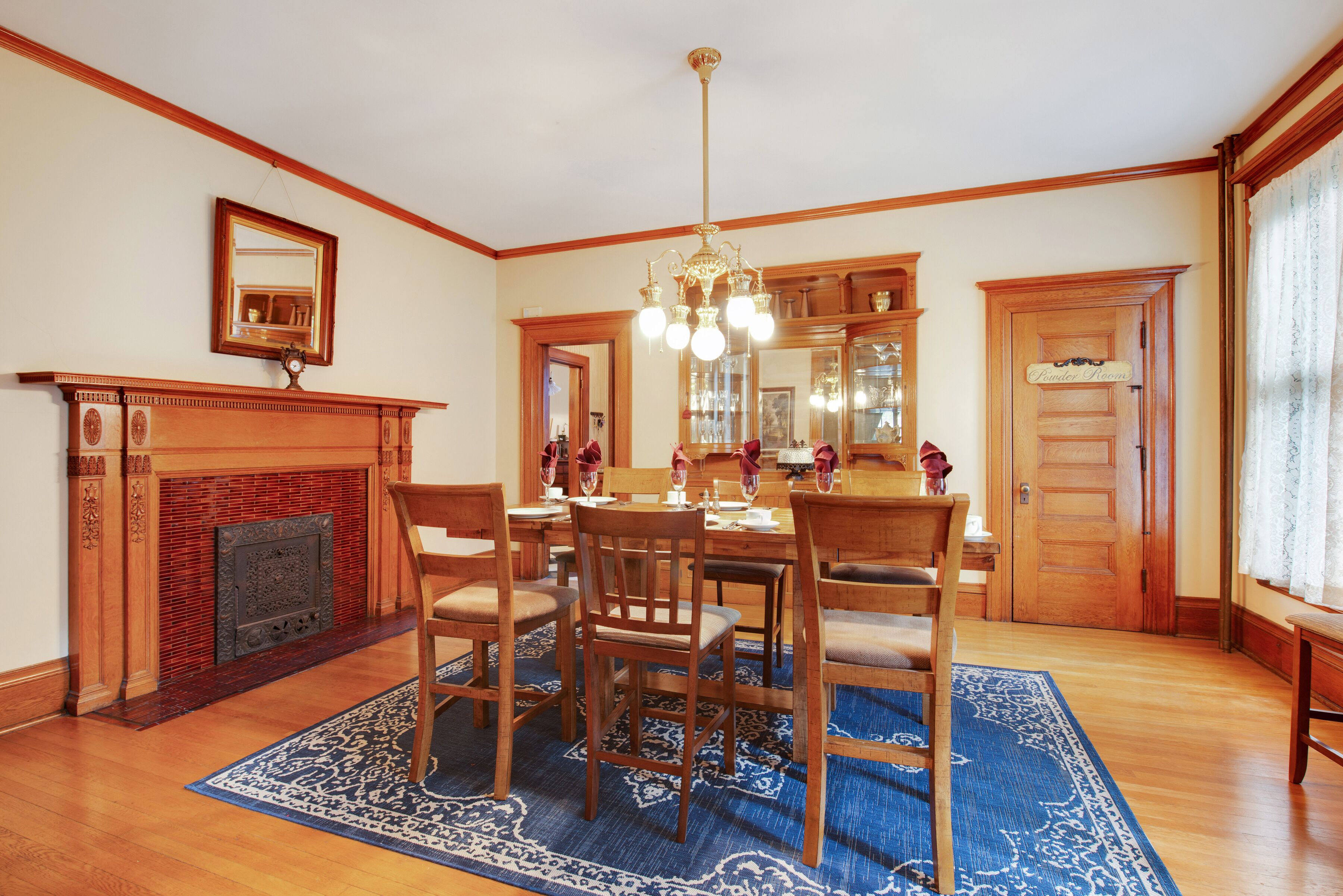 A dining room with a wooden table set for six, surrounded by chairs, a blue patterned rug, and a fireplace with a mirror above it.