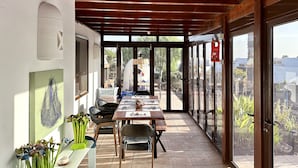 A covered outdoor dining area with a long table and chairs, large windows, and a view of the city.