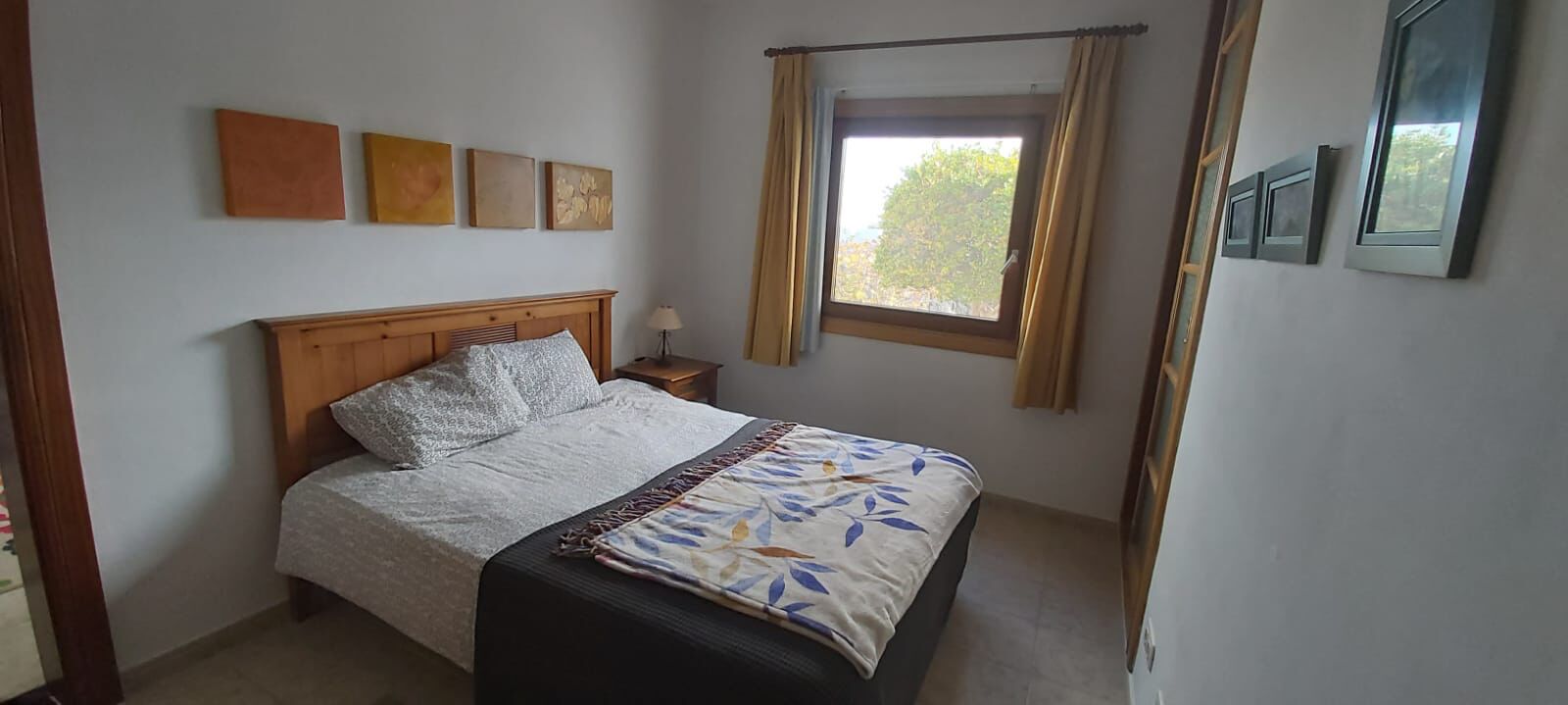 A bedroom with a bed, a nightstand, a window with curtains, and wall-mounted artwork.