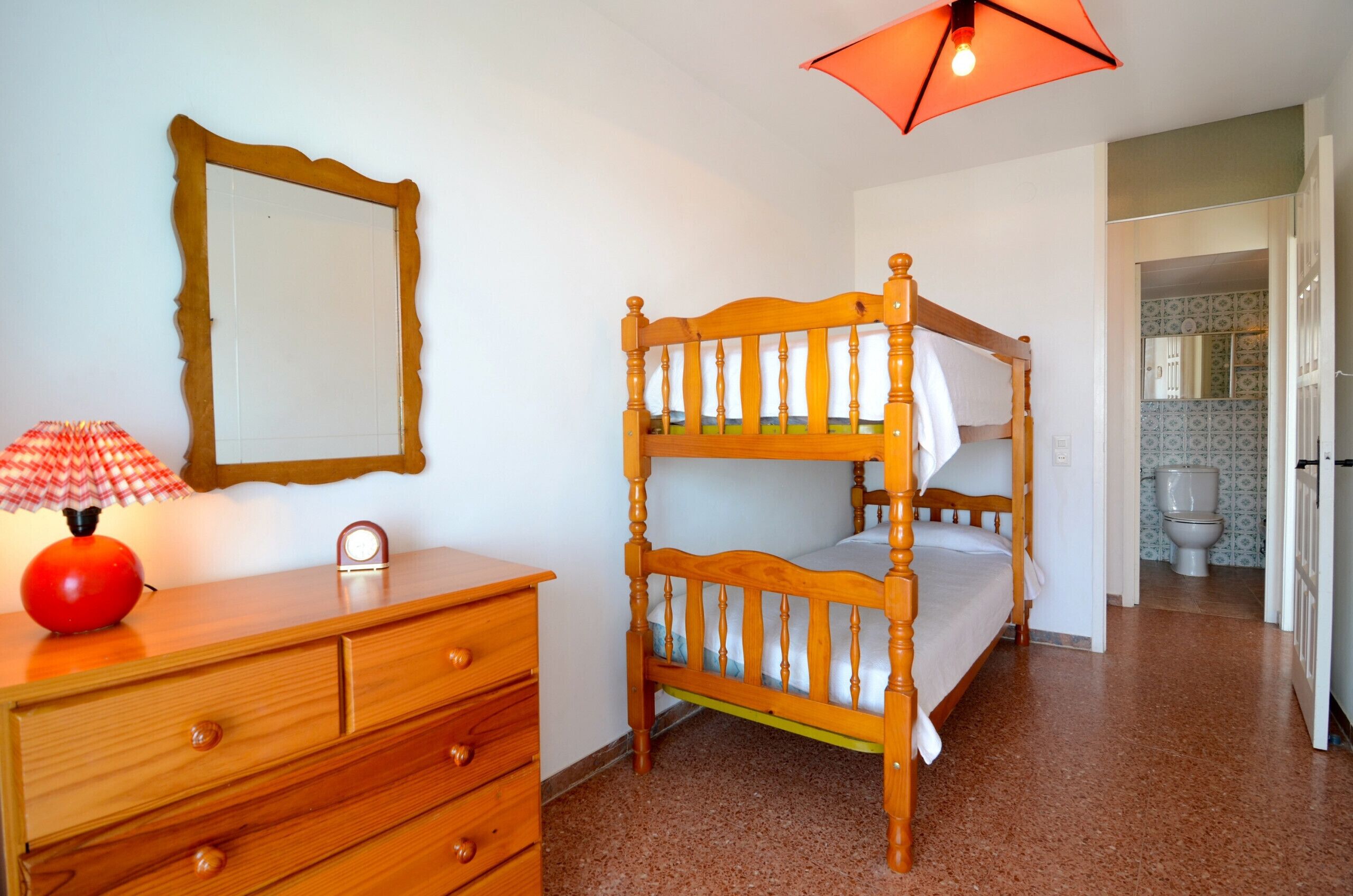 Un lit superposé avec une structure en bois, une commode en bois avec des tiroirs, un miroir et une lampe rouge.