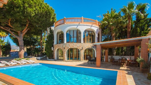 Vista a la piscina de una villa de estilo mediterráneo con fachada de piedra, ventanas grandes y un porche cubierto.