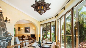 Un comedor con una larga mesa de madera dispuesta para cuatro personas, una chimenea de piedra y grandes ventanales que ofrecen vistas al exterior.