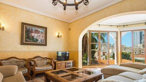 Una sala de estar con televisor, muebles de madera y un ventanal con vistas a edificios y palmeras.