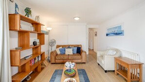 A living room with a brown leather sofa, a white armchair, wooden shelves, and a dining table with a bowl of cookies. - 2 Bed in Woolacombe (oc-beac4) (Woolacombe)