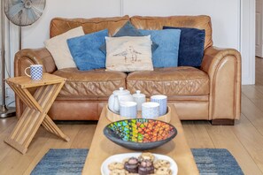A brown leather sofa with cushions, a small wooden side table with a mug, a tray with jars and a bowl of cookies, and a floor lamp. - 2 Bed in Woolacombe (oc-beac4) (Woolacombe)