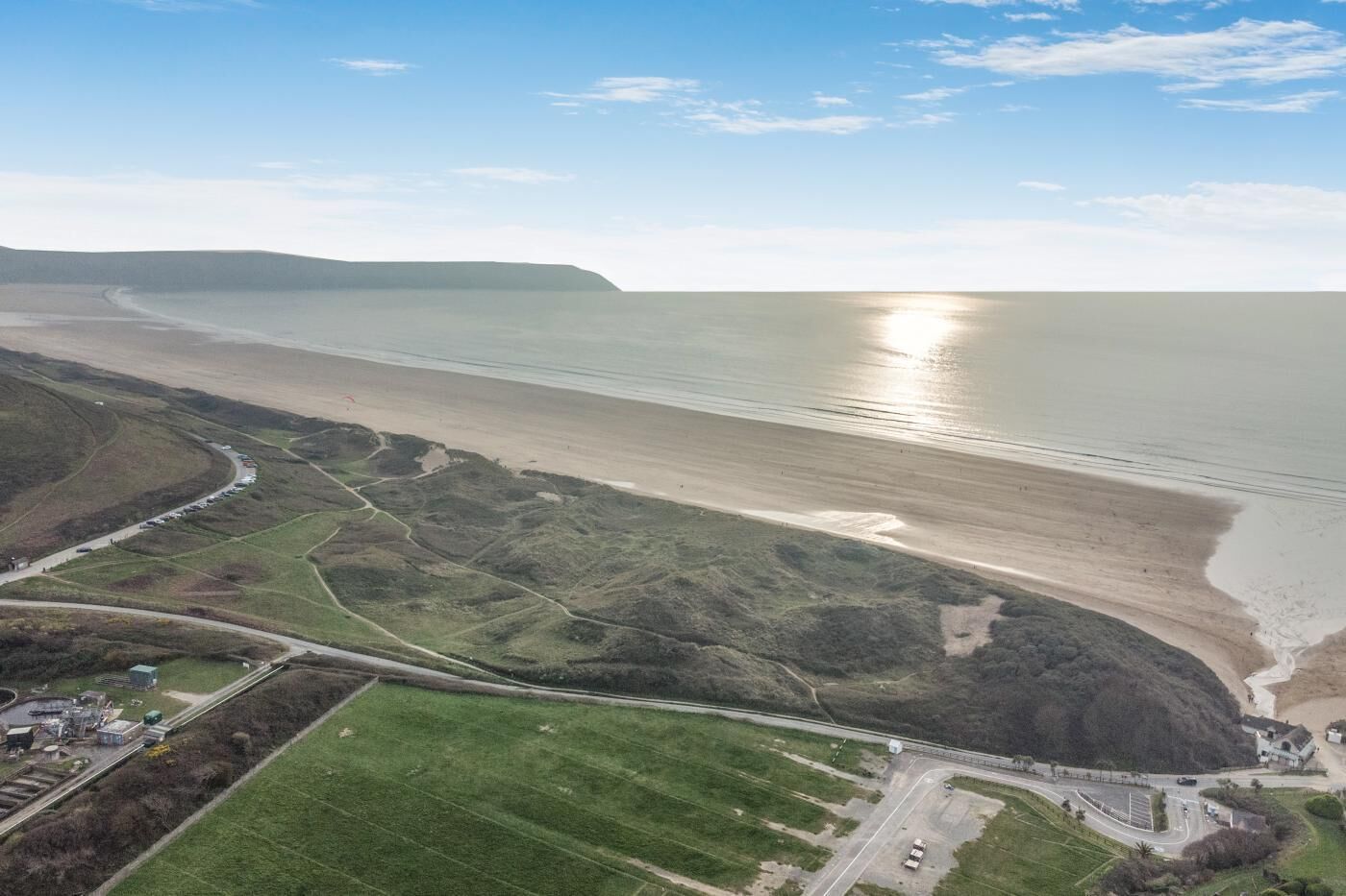 Vista aérea de una zona costera con playa, campos verdes y una carretera que conduce a un edificio.