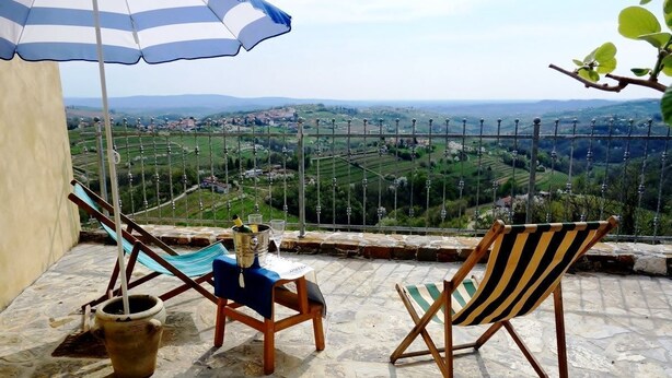 A terrace with a view of a hilly landscape, two wooden chairs, a striped umbrella, and a table with a metal bucket. - Little Vista, Smartno, Goriska Brda - 2 Bedroom Holiday House (Kojsko)