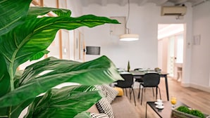 A modern living room with a large green plant, a black and white patterned sofa, a dining area with a table and chairs, and a kitchen visible in the background. - Born - Habitat Apartments (Barcelona)