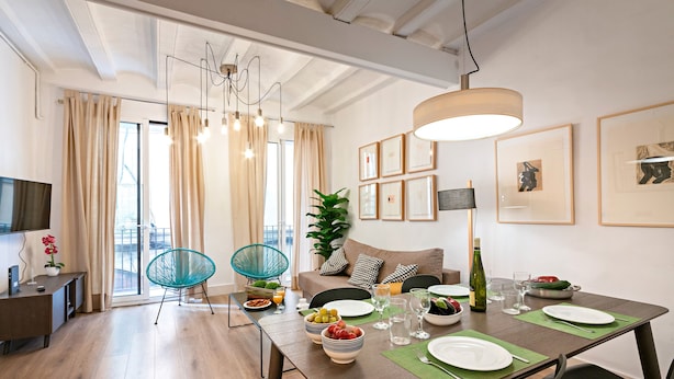 A modern dining area with a wooden table set for two, a sofa, and a television in the background. - Born - Habitat Apartments (Barcelona)