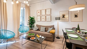 A modern living room with a sofa, two chairs, a dining table set for two, and a large window with curtains. - Born - Habitat Apartments (Barcelona)
