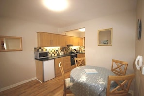 A compact kitchen with wooden cabinets, a microwave, and a small refrigerator. There is a dining table with chairs and a clock on the wall.