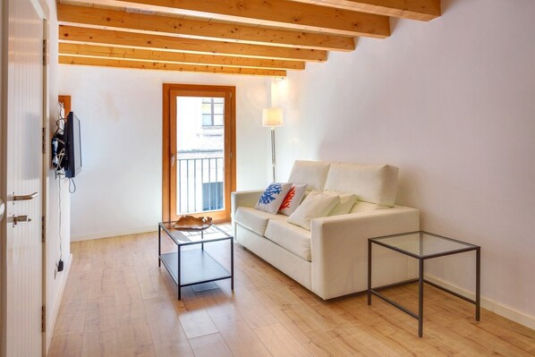 A living room with a white sofa, a glass coffee table, and a small side table. There is a wooden ceiling with exposed beams, a door leading outside, and a lamp on a floor lamp.