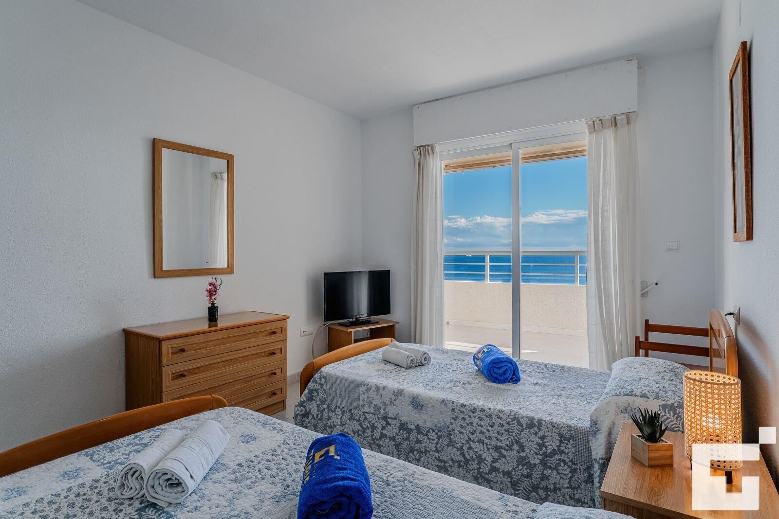 A hotel room with two beds, a TV, a dresser, and a view of the sea.