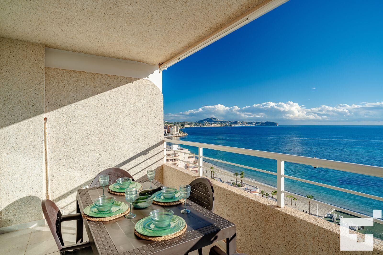 A balcony with a table set for dining, overlooking a coastal view with buildings and the sea.