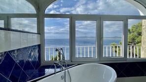 Een badkamer met een groot bad, uitzicht op zee en een balkon met een hekwerk.