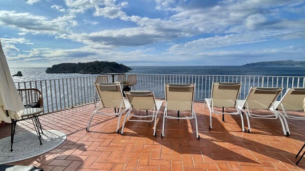 Een terras met zes loungestoelen en een tafel, met uitzicht op de kust, de zee en een eiland in de verte.