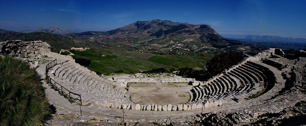 Un amphithéâtre antique doté de gradins en pierre et d’une scène centrale, le tout entouré d’un paysage montagneux.