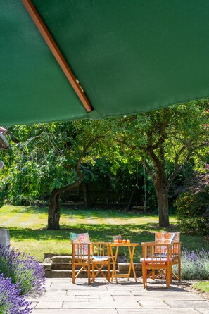 An outdoor seating area with two wooden chairs and a table, under a green canopy, surrounded by lush greenery and a stone pathway.