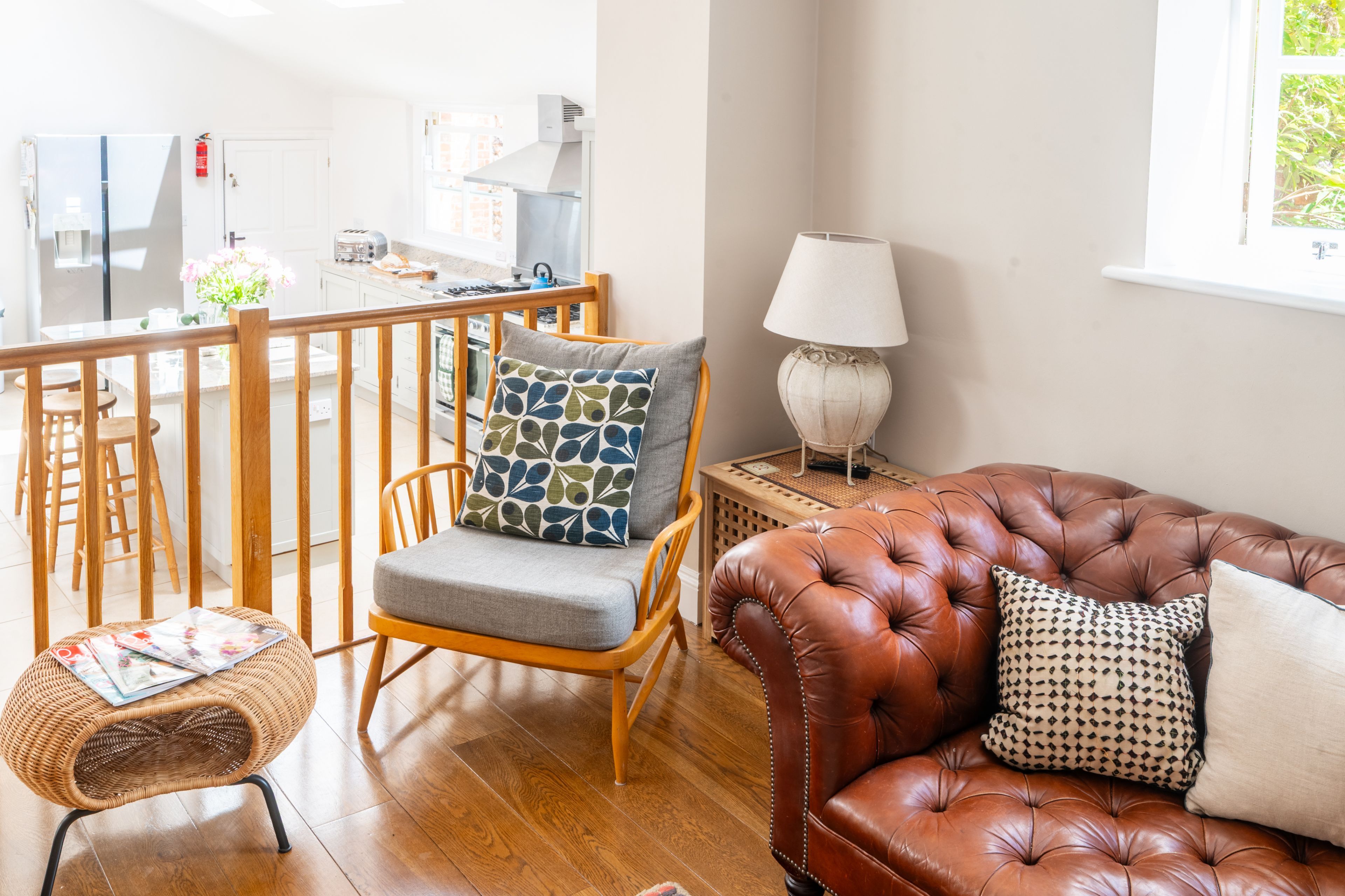 Un salon avec un canapé en cuir marron, un fauteuil en osier, une table à manger en bois et, au fond, une cuisine.