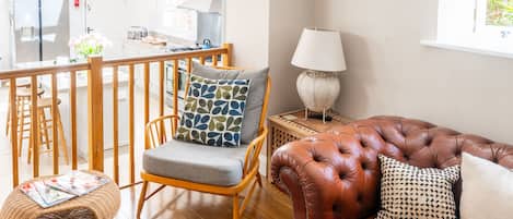A living room with a brown leather sofa, a wicker chair, a wooden dining table, and a kitchen in the background.