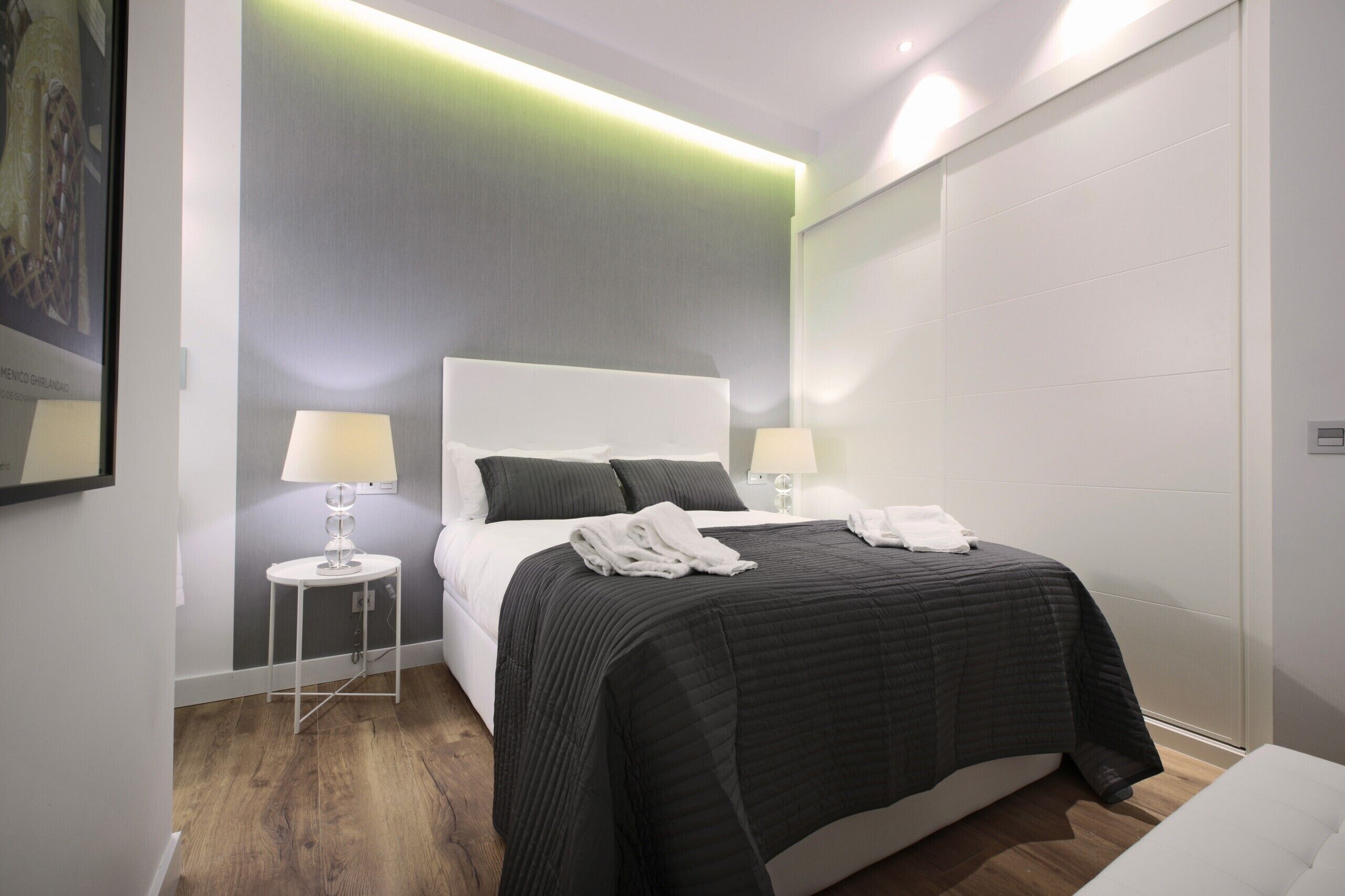 Una habitación de hotel moderna con una cama grande, dos mesitas de noche con lámparas, una pared de acento gris y suelo de madera.