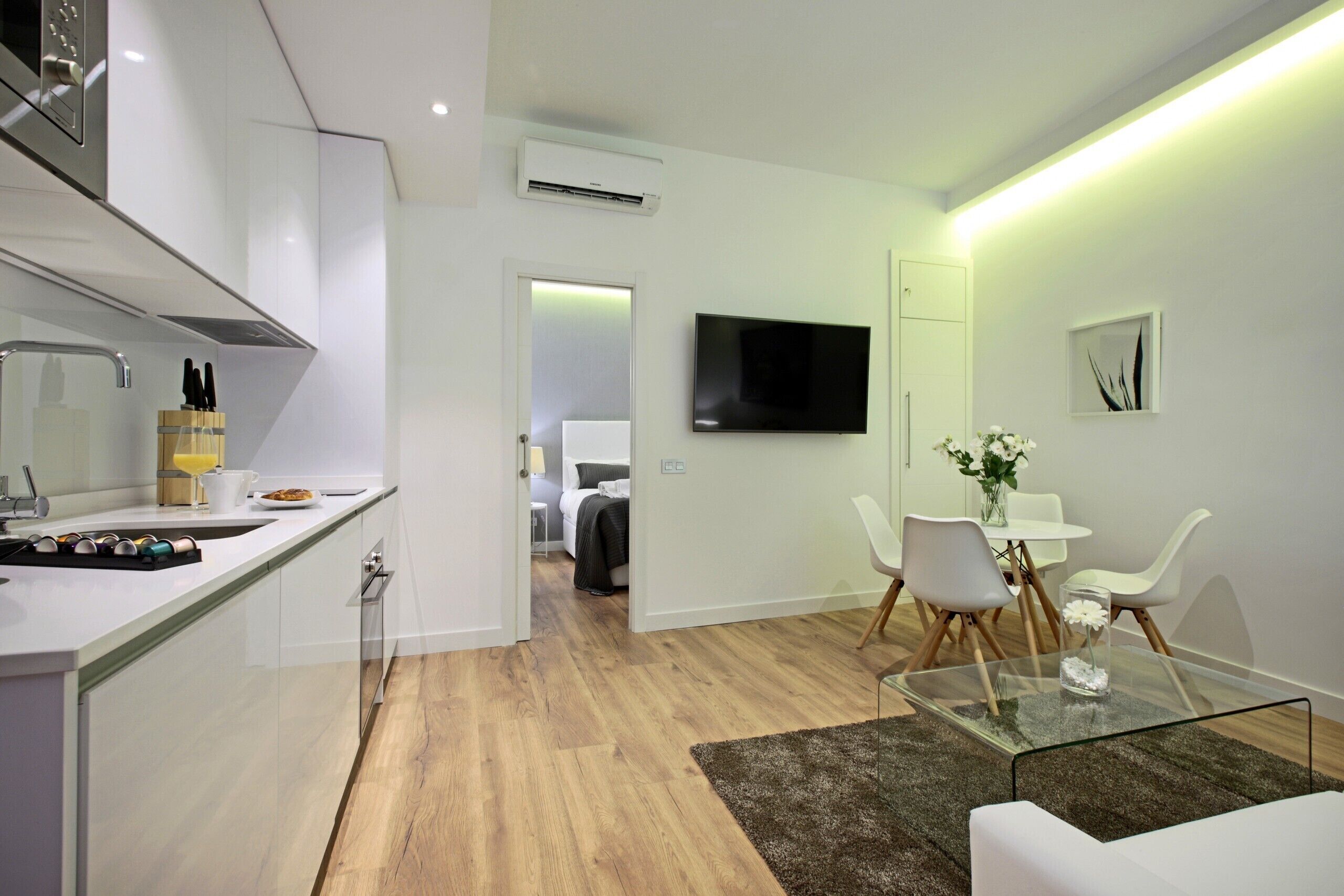Una cocina moderna con horno integrado, un comedor con mesa de vidrio y un espacio de sala con televisor de pantalla plana.
