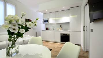 Cocina moderna con armarios blancos, microondas y horno empotrado. Cuenta con un comedor con mesa y sillas blancas, y un jarrón con flores blancas sobre la mesa.