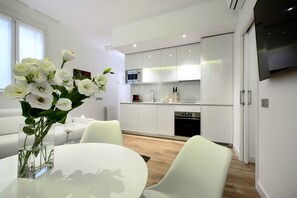A modern kitchen with white cabinets, a microwave, and a built-in oven. There is a dining area with a white table and chairs, and a vase of white flowers on the table.