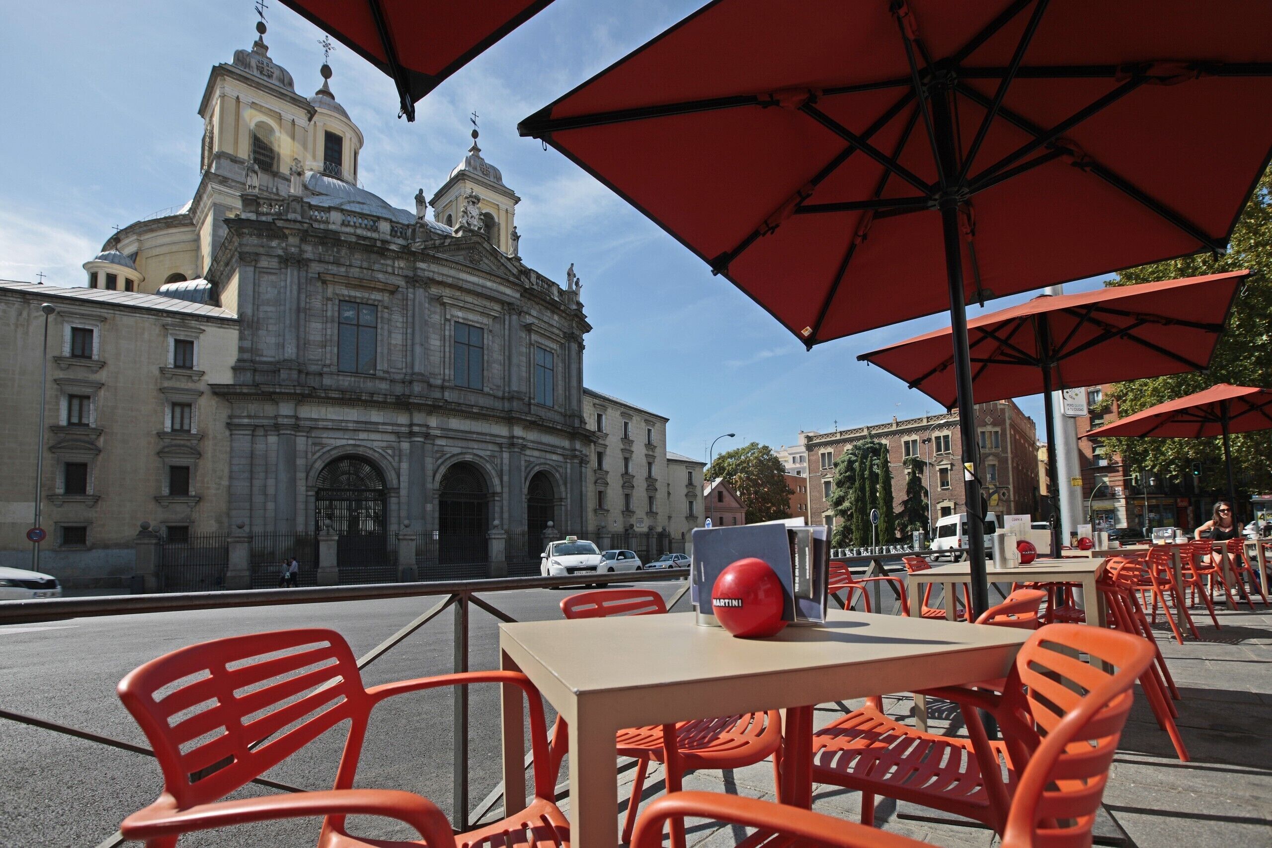 Mobiliario exterior con sombrillas rojas, una mesa con un objeto rojo y un edificio histórico al fondo.
