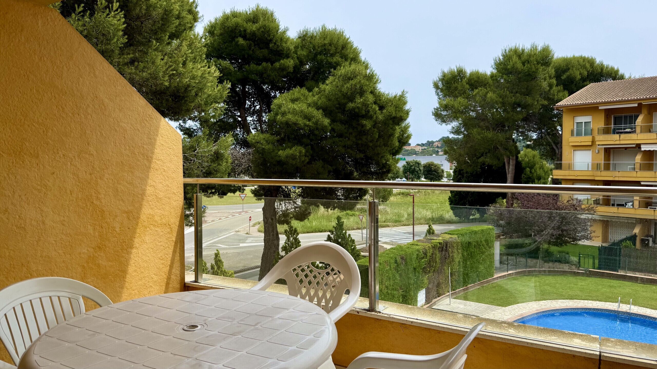 A balcony with a table and chairs, a pool, and a view of a road and trees.