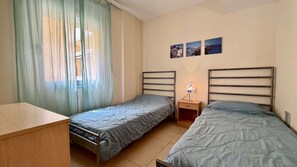 Ein Schlafzimmer mit zwei Einzelbetten, einem Nachttisch mit Lampe und einem Fenster mit durchsichtigen Vorhängen.