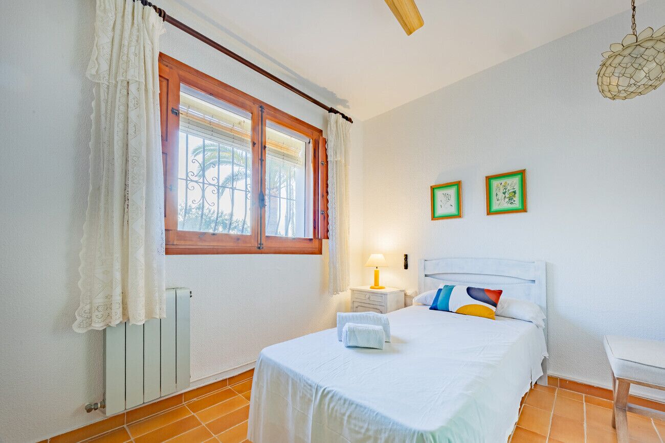 A bedroom with a bed, a nightstand, a lamp, a radiator, a window with curtains, and two framed pictures on the wall.