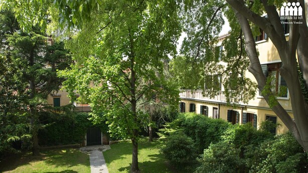 Un patio con un camino, abundante vegetación y un edificio con múltiples ventanas y balcones.
