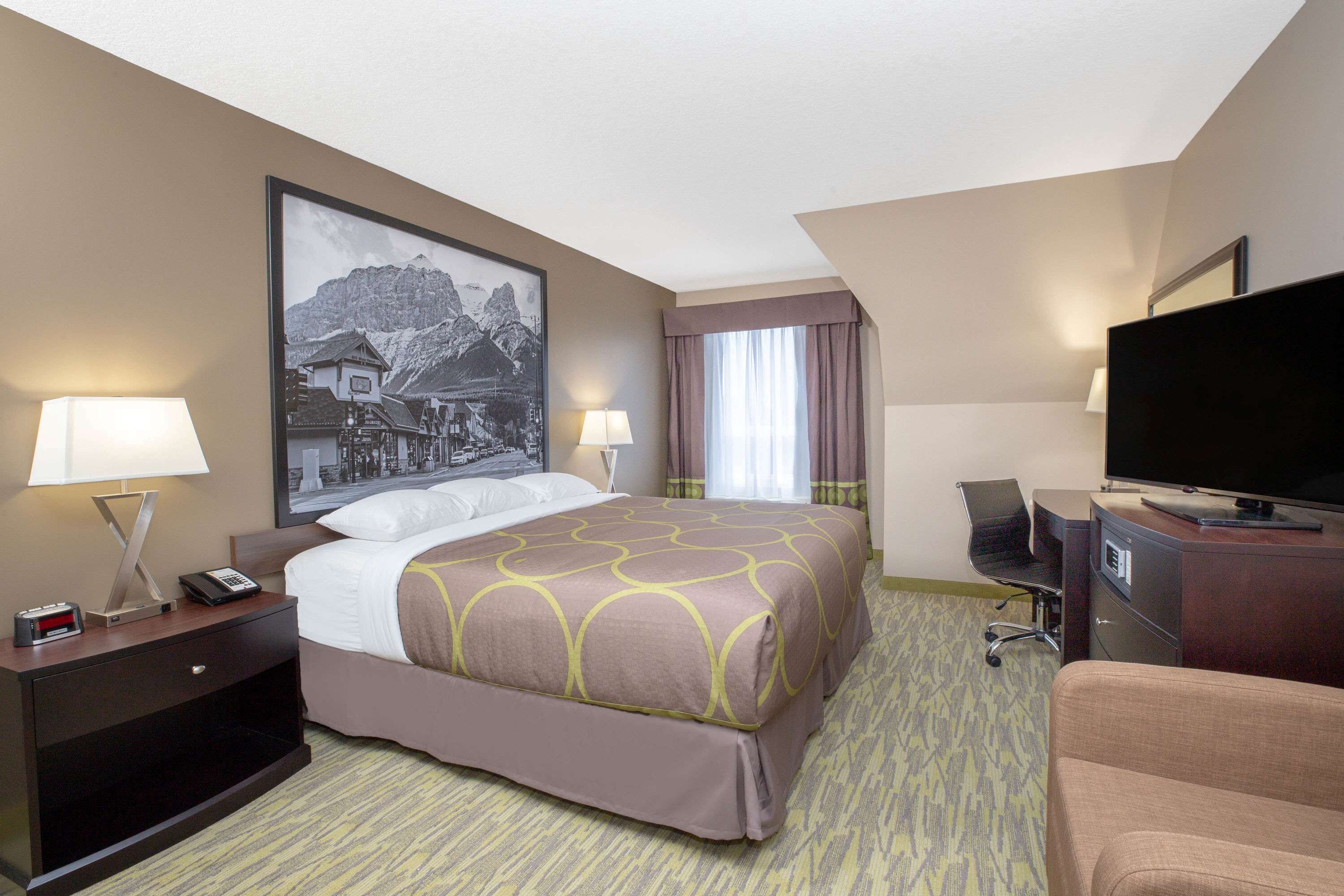 A hotel room with a bed, two bedside tables with lamps, a flat-screen TV, a desk with a chair, and a large framed picture on the wall.
