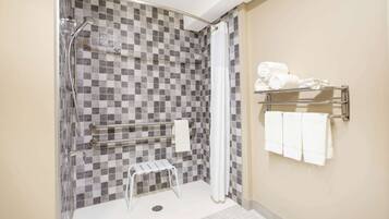 A bathroom with a checkered tile wall, a shower with a grab bar, a white towel rack, and a small step stool.