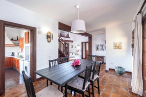 Un comedor con una mesa de madera oscura, sillas, una lámpara colgante y una cocina visible a través de un vano abierto.