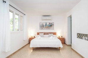 Un dormitorio con una cama grande, una mesita de noche, un aire acondicionado de pared y una ventana con cortinas.