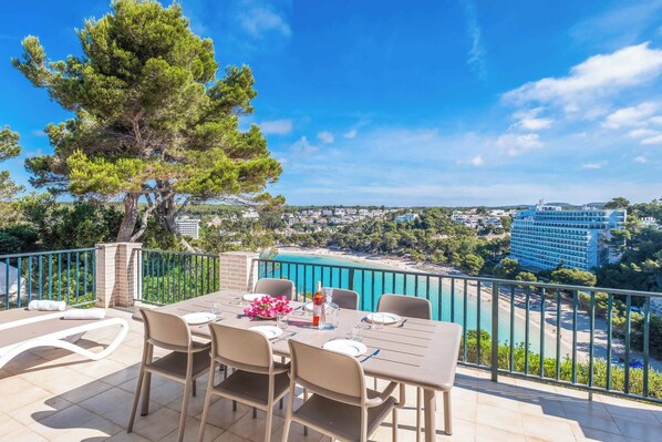 An outdoor dining area with a table set for two, overlooking a coastal view with a pool and buildings.